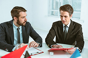 Zwei in schwarze Businessanzügen gekleidete, junge Männer unterhalten sich über ein Dokument auf einem roten Klemmbrett