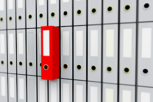 Eine Wand voller hellgrauer Ordner, aus welchem mittig ein roter Ordner griffbereit herausragt